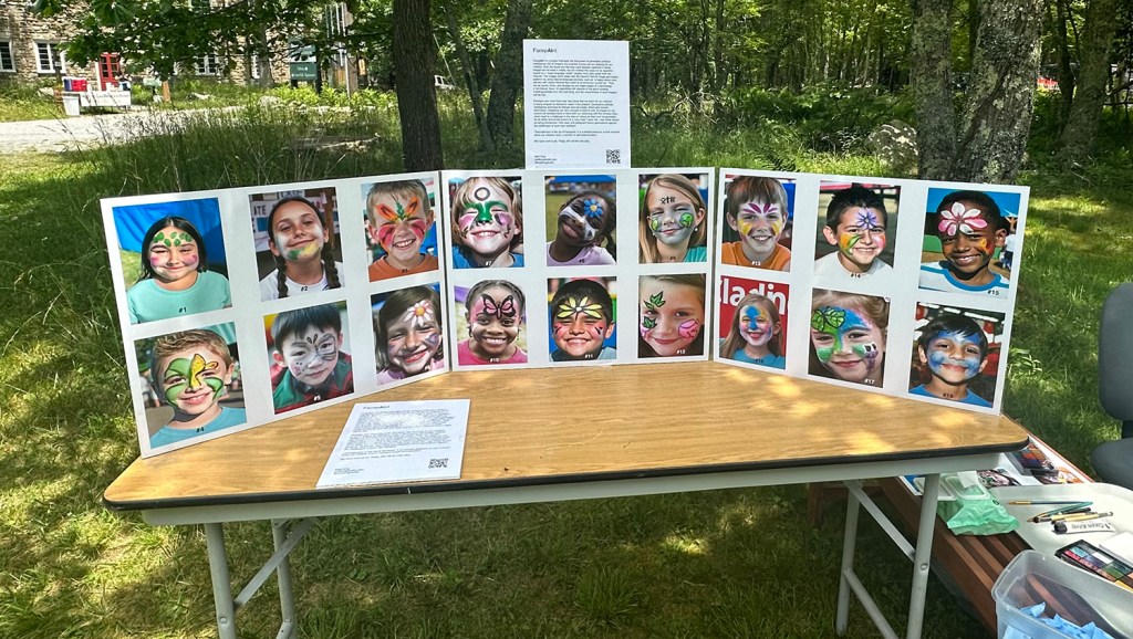Board of AI-generated face-paint designs (“flash”) on the fair table for AI Face Paint, 2023, by Matt King. Participatory face-painting performance.