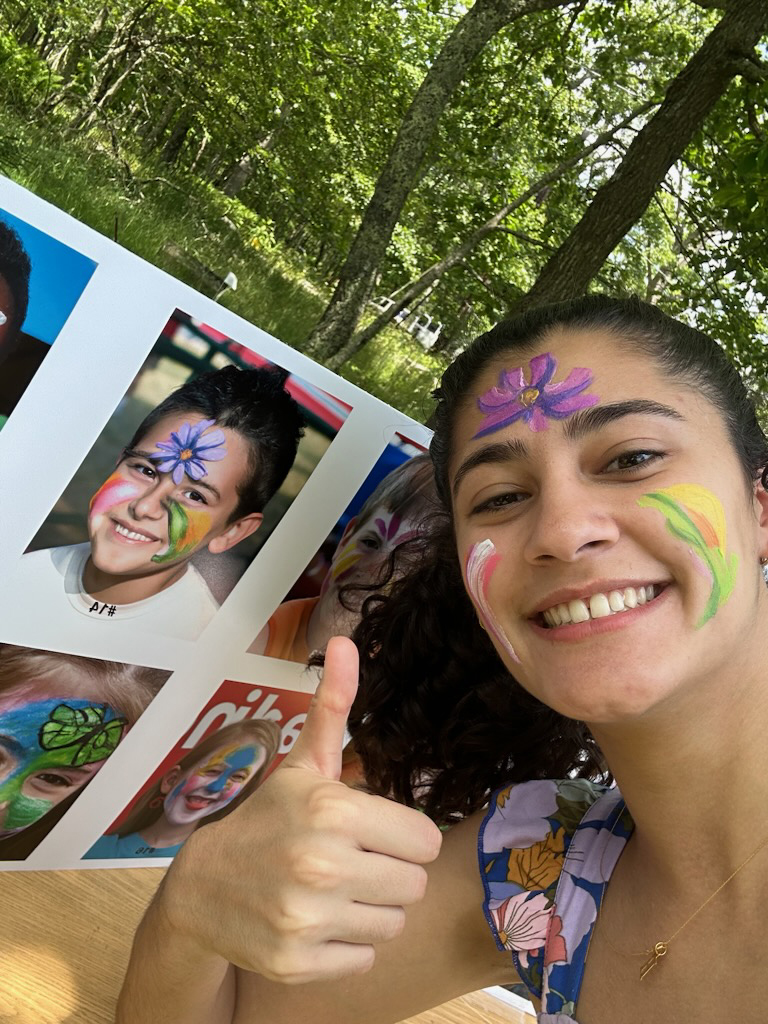 A smiling young woman gives a thumbs up in front of a display board featuring several children with colorful face paint designs, including flowers and nature themes, in a green outdoor setting.