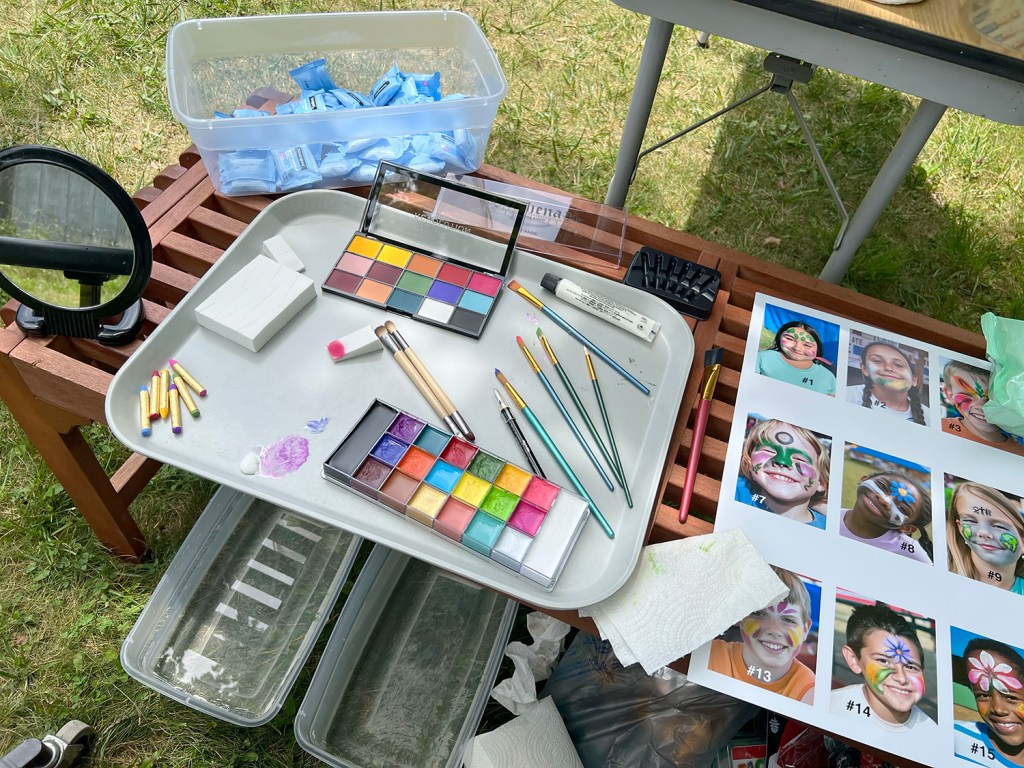 Face-painting table with paints, brushes, and printed designs for AI Face Paint, 2023, by Matt King. Participatory face-painting performance.